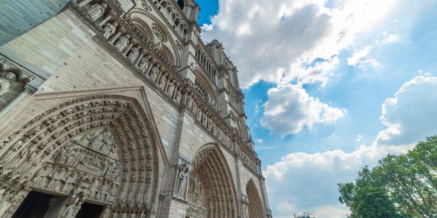 Low-angle view of the Notre-Dame Cathedral in Paris, showcasing its intricate stone carvings and Gothic architecture against a bright blue sky with fluffy clouds.