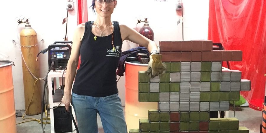 Welder Shanen Aranmor stands beside a pixel-art sculpture welded from painted steel blocks at her Weld Like a Girl shop in Yuma, Arizona.
